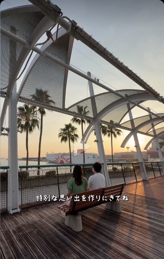 夕日の中、海辺のベンチに並んで座るふたり。奥にさんふらわあが見える