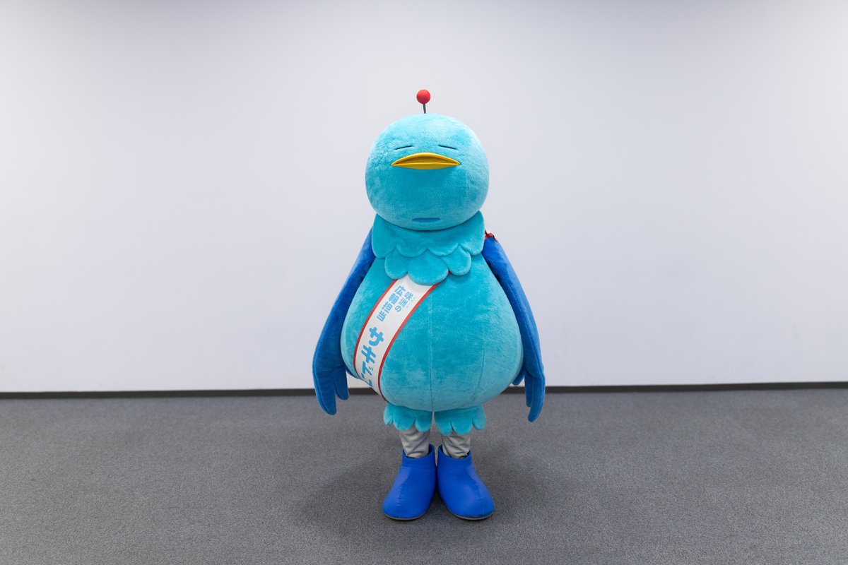 白い壁の前に立つ、水色の鳥のキャラクター、サキドリちゃん